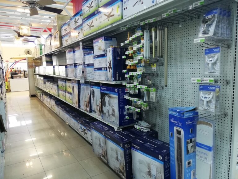 Aisle in a hardware store on Roatan, Honduras, featuring ceiling fans, window fans, and other appliances on metal shelves.