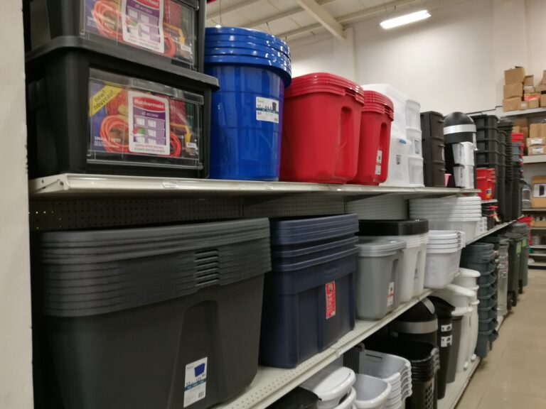 Stacks of plastic storage bins and buckets in various colors are displayed on metal shelving in a hardware or home improvement store in Roatan, Honduras.