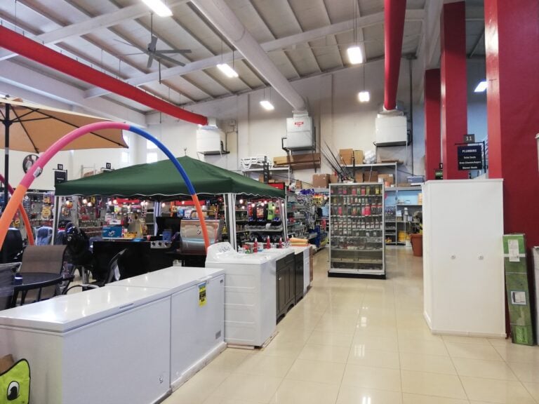 The interior of a hardware or home improvement store in Roatan, Honduras, featuring laundry appliances, shelving stocked with tools and supplies, and signage indicating plumbing and other department sections.