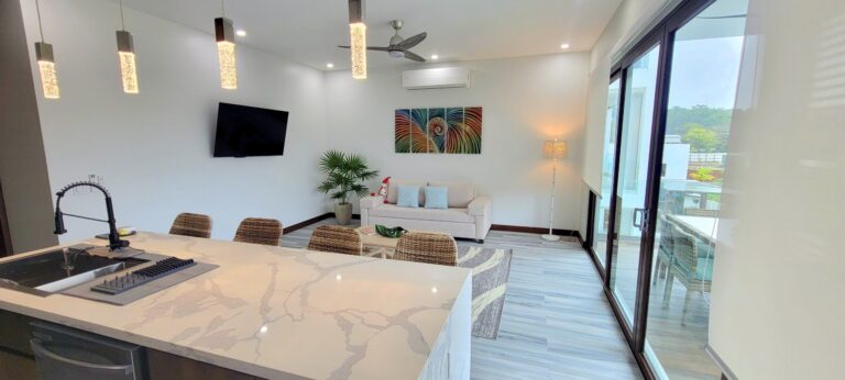 Modern living room in a Caribbean-style apartment on Roatan, Bay Islands, Honduras, featuring a white sofa, colorful wall art, a large flat-screen TV, and a balcony with outdoor seating overlooking lush greenery.
