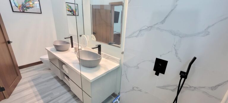 Modern bathroom with a double vanity featuring two vessel sinks, black faucets, and a large mirror, set against white marble walls with gray veining in Roatan, Honduras.