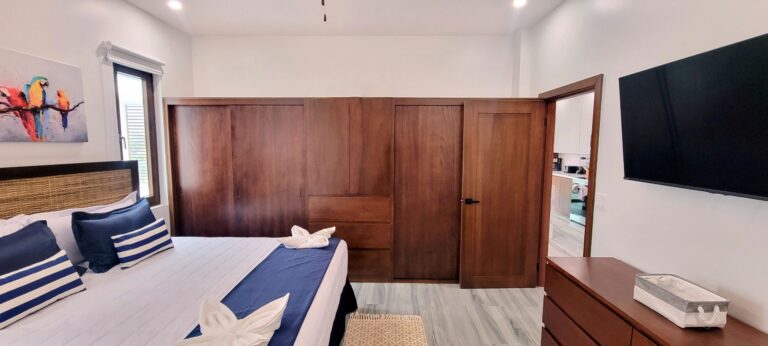 A bedroom in a Roatan holiday rental featuring a bed with navy and white striped pillows, a rattan headboard, a wooden wardrobe, and a wall-mounted flat-screen TV with a view into the kitchen area in the background, set against white walls and light grey wood flooring in Honduras.