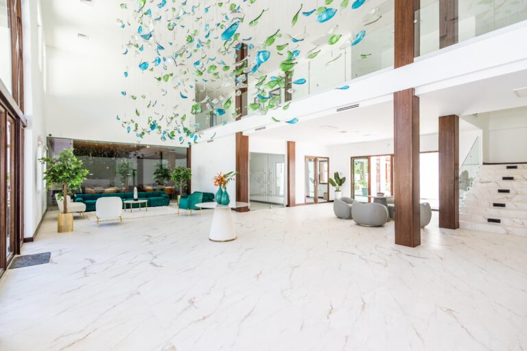 Interior view of a modern, spacious lobby in Roatan, Honduras, featuring white marble flooring, contemporary seating with teal and white furniture, large glass doors, and colorful glass art hanging from the ceiling.