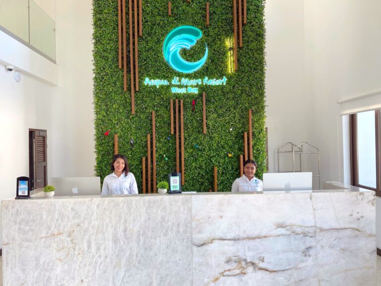 Reception area at Acqua di Mare Resort in West Bay, Roatan, Honduras, featuring a green vertical garden wall with a neon wave sign and two smiling staff members behind a white marble counter with Apple computers.