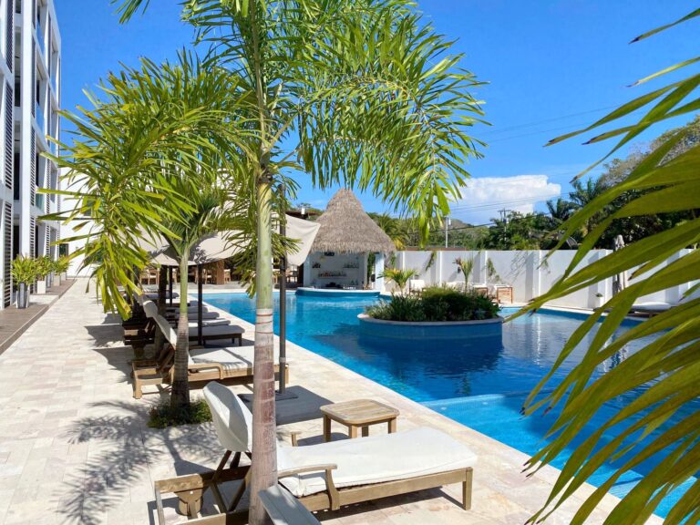 Tall green palm trees situated near a swimming pool surrounded by beige tiles at a business on Roatan, Honduras, with a thatched-roof cabana in the background and a modern white building nearby under a clear blue sky.