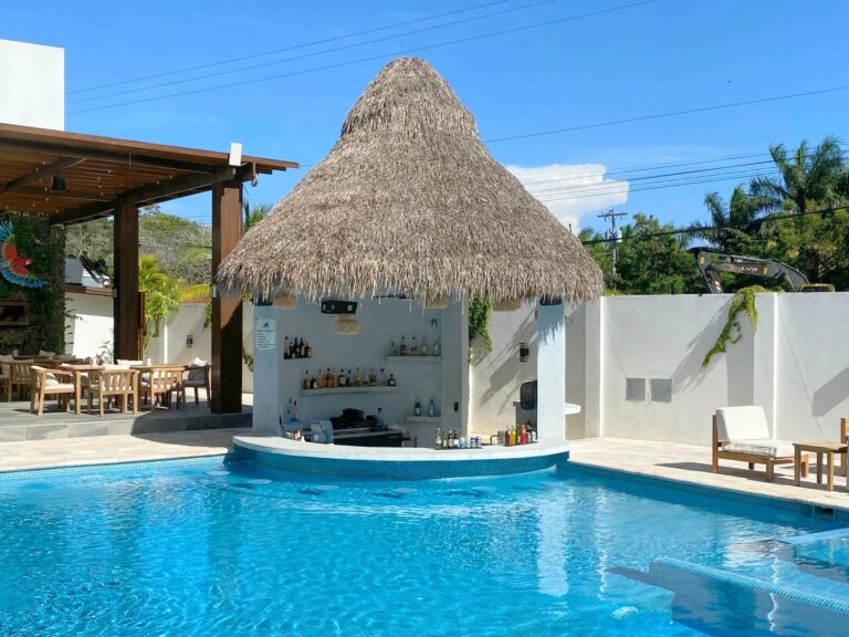 A vibrant swimming pool with clear blue water, surrounded by lounge chairs and a tiki hut bar made of wood and thatch on Roatan, Honduras, under a bright blue sky.