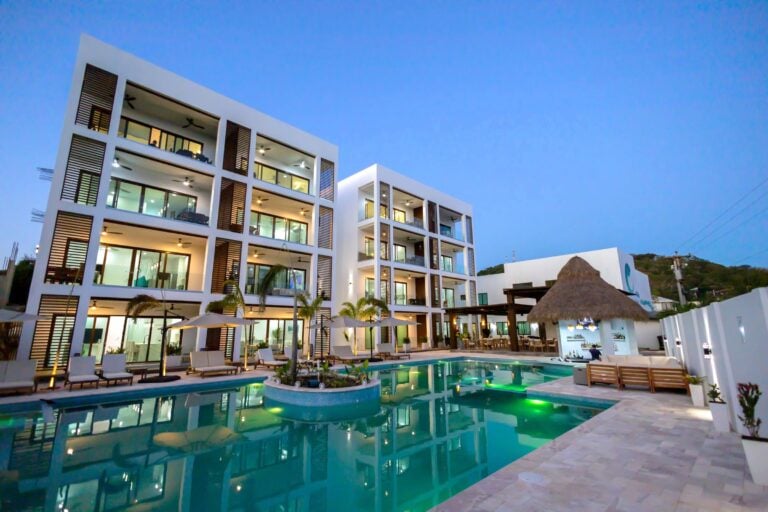 Modern white hotel building with multiple floors and balconies overlooking a swimming pool, surrounded by palm trees and tropical landscaping on Roatan, Honduras during evening hours.