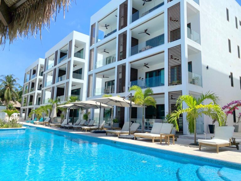 View of a modern white building with multiple balconies and glass railings on Roatan, Honduras, featuring a swimming pool in the foreground surrounded by lounge chairs and umbrellas under clear blue skies.