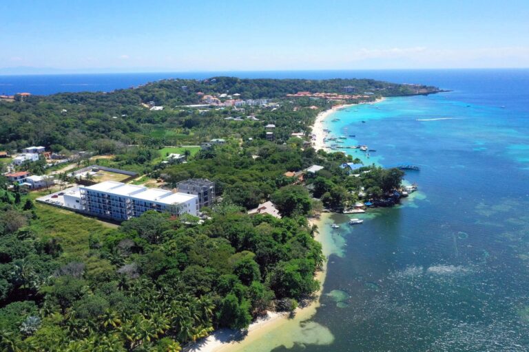 A panoramic aerial view of Roatan, Honduras, showing lush green hills, a sandy beach with boats anchored in the turquoise waters, and residential and hotel buildings along the coastline under a clear blue sky.
