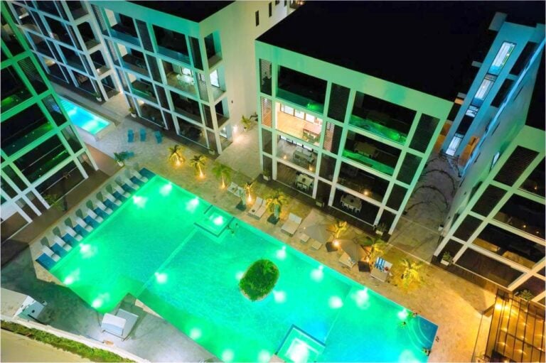 A nighttime aerial view of a modern hotel in Roatan, Honduras, featuring a brightly illuminated swimming pool surrounded by palm trees and sun loungers, with multiple balconies overlooking the pool area.