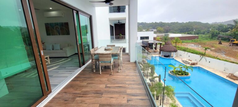 Modern rooftop balcony in Roatan, Honduras, overlooking a swimming pool with a thatched-roof tiki hut and lush tropical landscaping, with construction equipment and hills in the background.