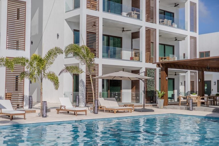 A modern white apartment building in Roatan, Honduras features multiple balconies with glass railings, wooden shutters, ceiling fans, and an outdoor pool area with lounge chairs, umbrellas, and tropical palm trees.