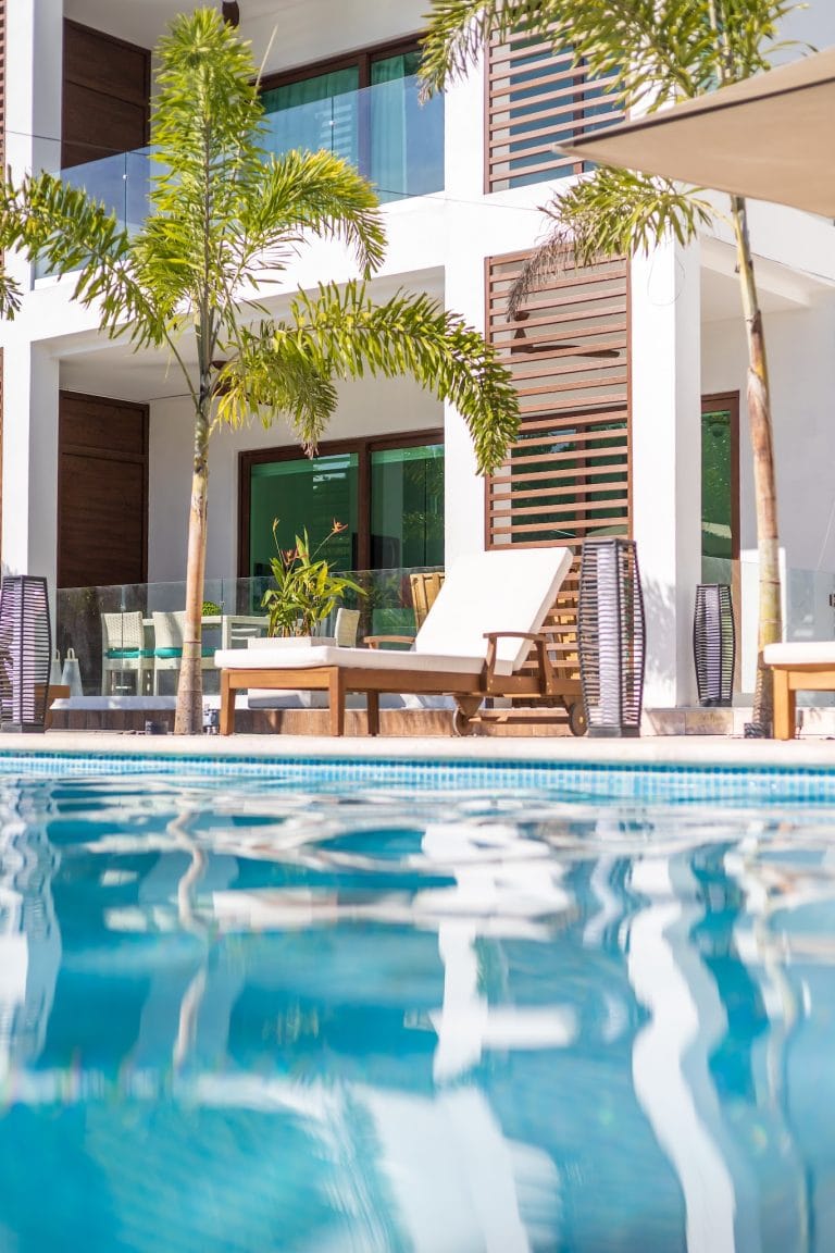 A modern multi-story building with white walls, wooden accents, and glass balconies surrounded by palm trees and lounge chairs near a swimming pool in Roatan, Honduras.