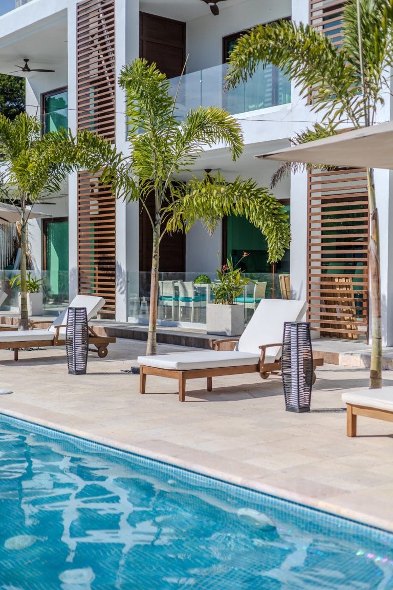 A swimming pool area at a modern hotel or resort in Roatan, Honduras, featuring lounge chairs, palm trees, umbrellas, and a multi-story building with balconies and wooden accents.