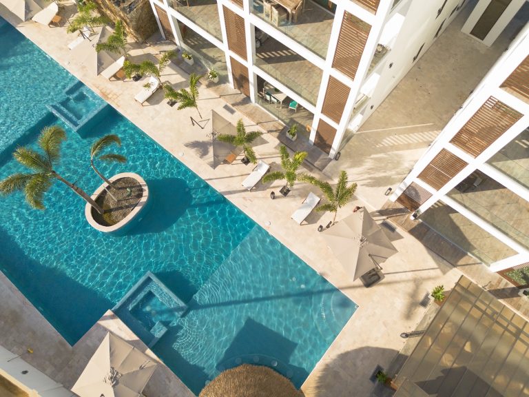 Aerial view of a modern building with balconies and wooden shutters overlooking a large, curved swimming pool with palm trees and lounge chairs along the poolside in Roatan, Honduras.