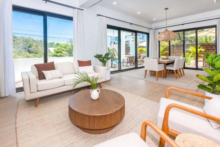 Bright and airy living room in a modern home on Roatan, Honduras, featuring large windows, white furniture, and indoor plants with a view of outdoor patio area.