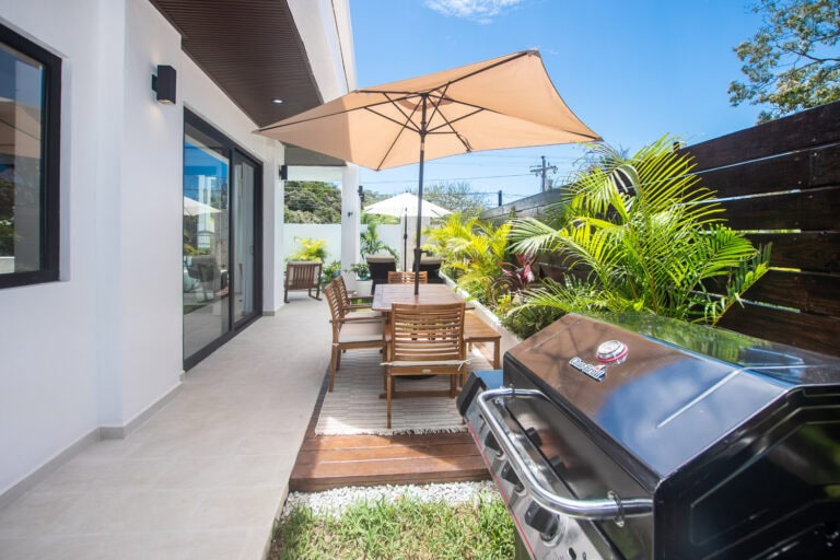 A modern outdoor patio in Roatan, Honduras, with a wooden dining table, chairs, large umbrellas, a gas grill, and lush tropical plants along a wooden privacy fence under a clear blue sky.