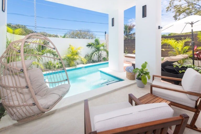 A modern outdoor patio in Roatan, Bay Islands, Honduras, featuring a small swimming pool, tropical plants, and comfortable seating with a hanging egg chair and lounge chairs under a white umbrella.