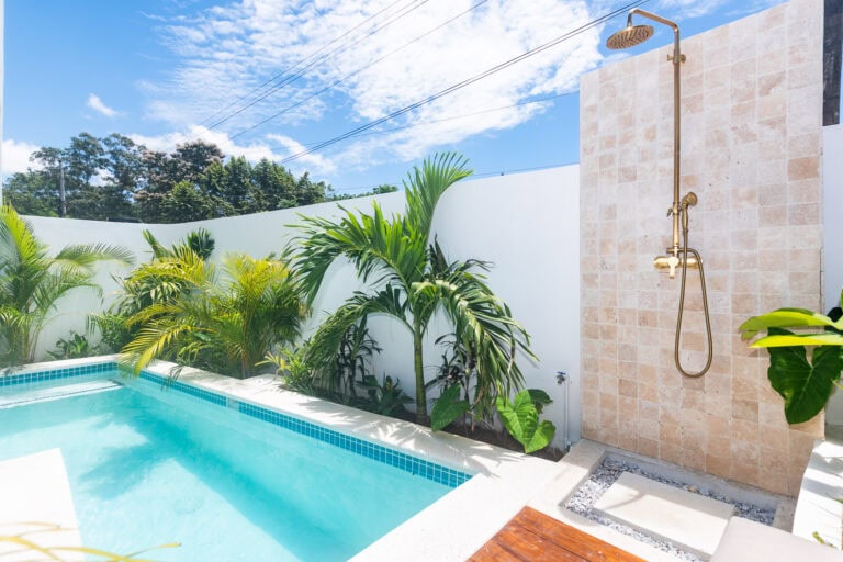 Relaxing outdoor private pool area with lush tropical plants and an outdoor shower in Roatan, Honduras under a bright blue sky with scattered clouds.