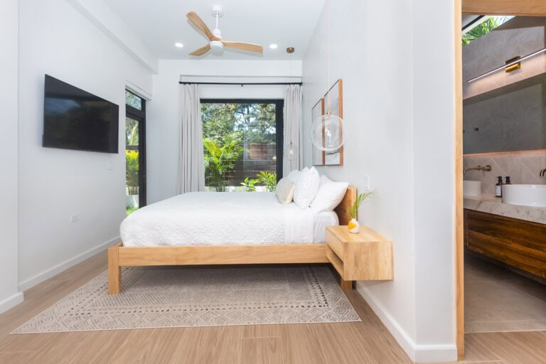 Modern bedroom with a wooden bed frame, white bedding, and large windows overlooking lush greenery in Roatan, Honduras.