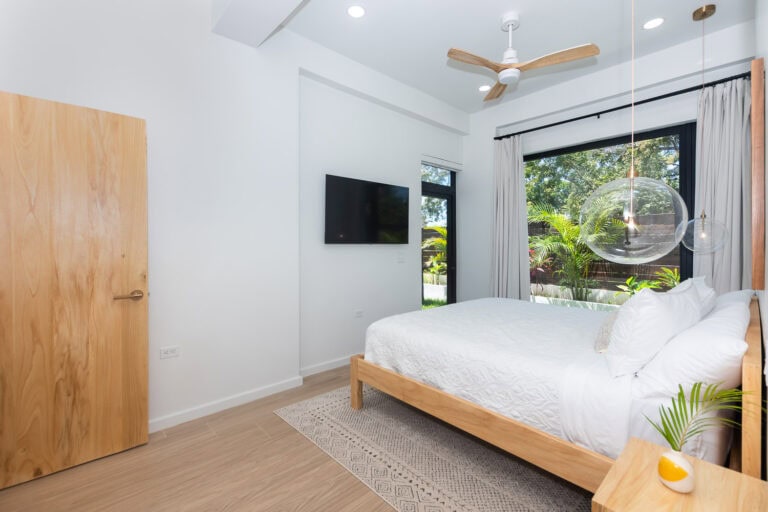 Modern bedroom in a tropical villa on Roatan with large windows overlooking lush greenery and a wooden door.