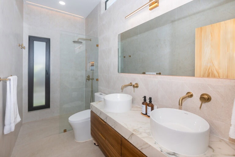 Modern bathroom in a luxury hotel on Roatan with dual vessel sinks, brass fixtures, a large mirror, and a walk-in glass shower with a rainfall showerhead.