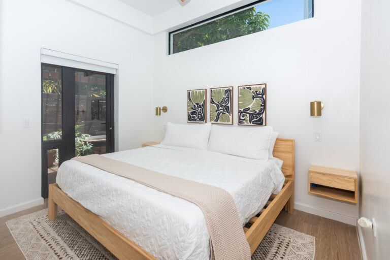Modern bedroom in a coastal home on Roatan with white walls, a wooden bed frame, and tropical artwork above the bed while large windows and a glass door provide natural light and views of lush greenery in Honduras.