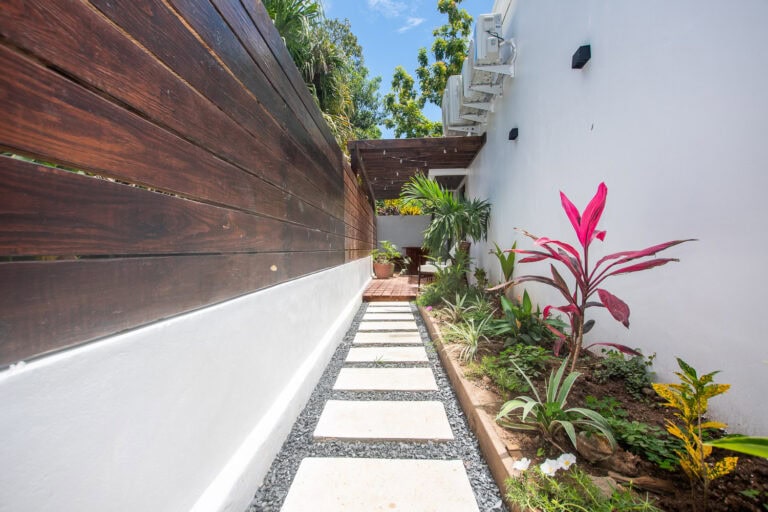 A narrow garden pathway with white stepping stones and black gravel, bordered by a white wall on one side and a lush tropical garden on the other, leading to an outdoor seating area on Roatan, Honduras.