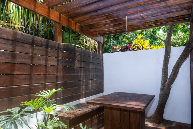 A cozy outdoor seating area in Roatan with a wooden pergola, white wall, lush tropical plants, and a small tree growing beside the bench