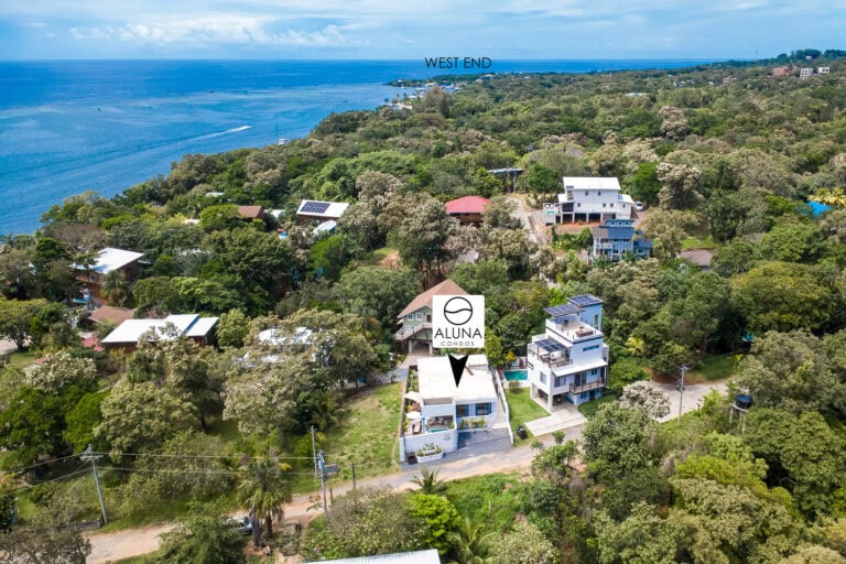 Aerial view of residential buildings and lush green trees along the coast of Roatan, Honduras, with the ocean in the background and the label 'West End' marking the area.