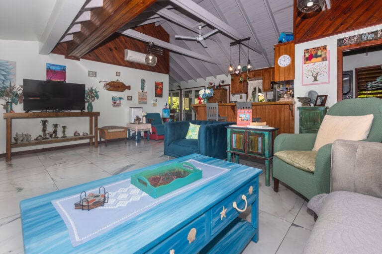 Living room with white walls, wooden ceiling beams, and colorful coastal decor in Roatan Honduras, featuring a television, paintings, and comfortable seating.