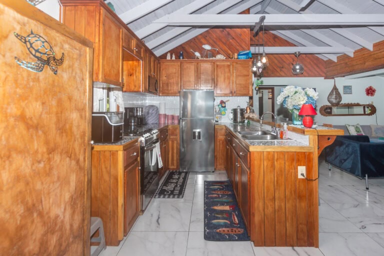 The image shows a cozy kitchen in a home on Roatan, Bay Islands, Honduras, featuring wooden cabinets, a stainless steel refrigerator, and marble countertops with a backyard view through a window.