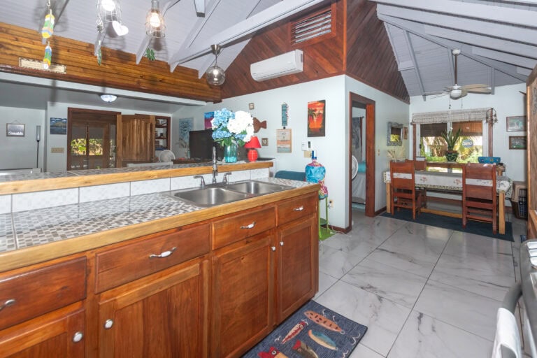 Interior view of a cozy Roatan home kitchen with wooden cabinets, a tiled island counter, and decorative hanging lights, overlooking a dining area with a wooden table and chairs, and lush greenery visible through the windows in Honduras.