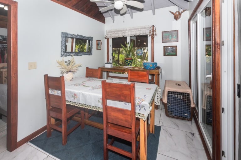 Interior view of a dining area in a house on Roatan, Honduras, featuring a wooden dining table with floral tablecloth, surrounded by matching wooden chairs, and a large window overlooking lush greenery outside.