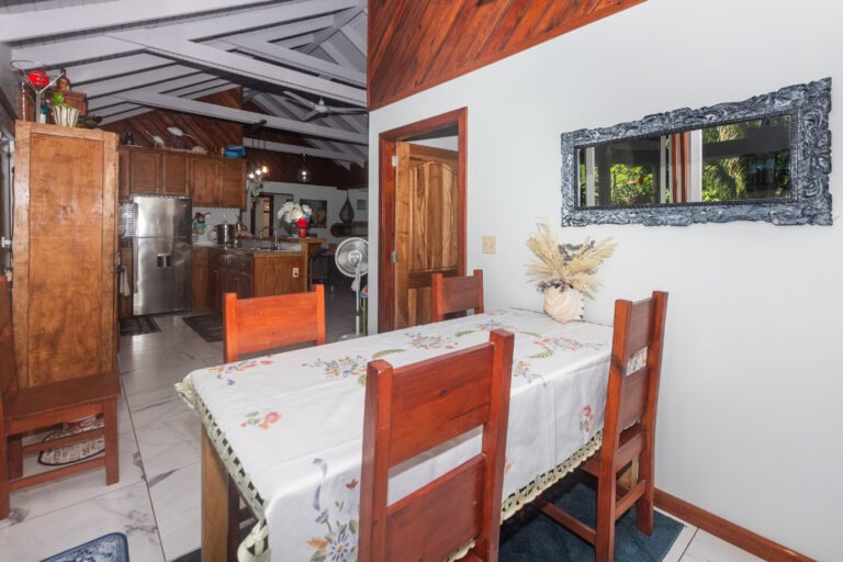 Interior view of a cozy dining area in Roatan, Honduras featuring wooden furniture, a floral tablecloth, and a large decorative mirror on a white wall with greenery reflected in it.