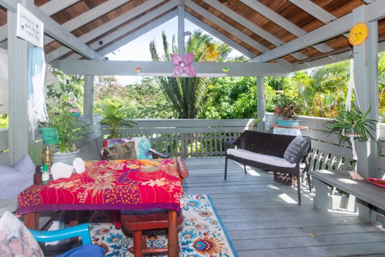 A cozy outdoor seating area on a wooden deck in Roatan, Honduras, decorated with colorful pillows, plants, and ocean-themed ornaments under a sloped roof with tropical greenery visible in the background.