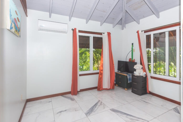 A bright living room in a house on Roatan, Honduras, featuring white walls, large windows with orange and white curtains, and a view of lush tropical greenery outside.