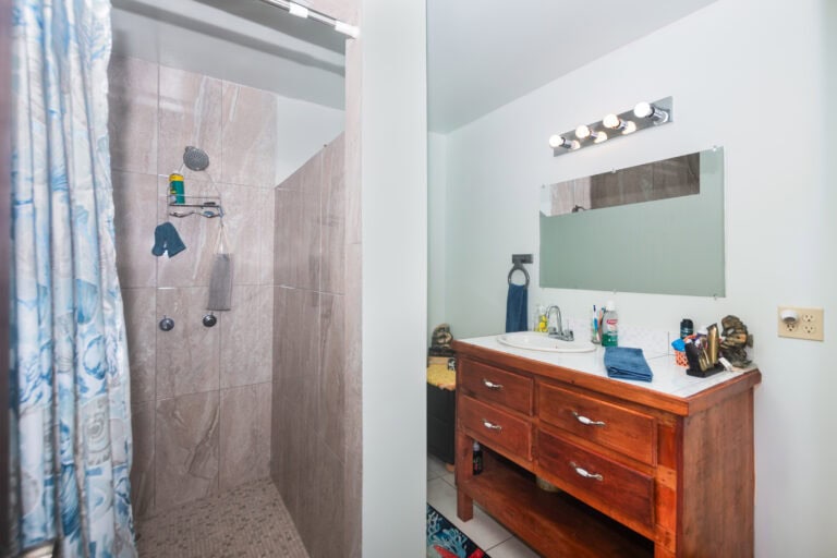 A bathroom in Roatan, Honduras featuring a shower area with beige tiles, a showerhead, and a blue and white patterned shower curtain, alongside a wooden vanity with a white sink, a large mirror, and various toiletries on the counter.