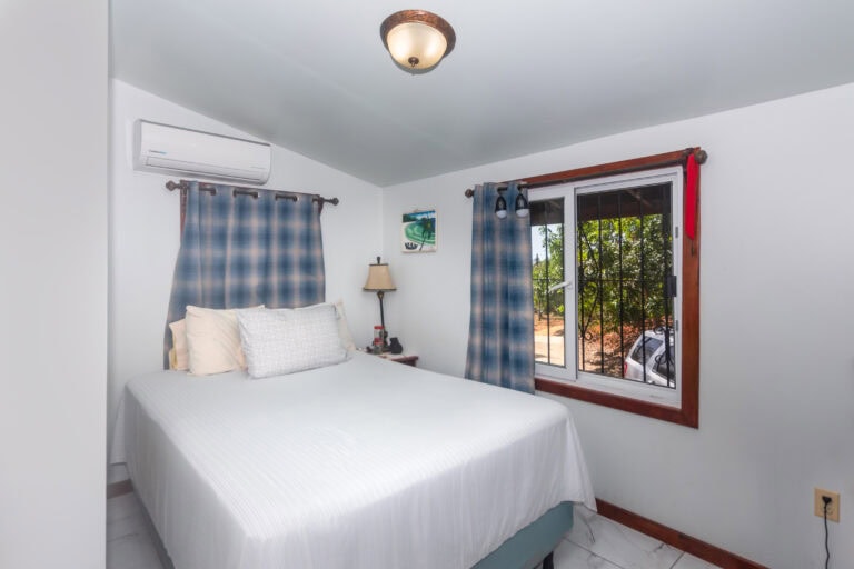 A clean, well-lit bedroom on Roatan with white walls, a large bed with white bedding, blue plaid curtains, and a window showing green tropical foliage outside in Honduras.