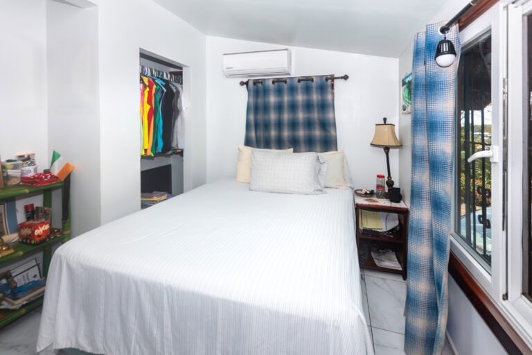 A cozy bedroom in Roatan with white walls, a large bed with white linens, and a closet filled with colorful clothes, complemented by a small bookshelf, a nightstand with a lamp, and a window with blue checkered curtains overlooking lush greenery.
