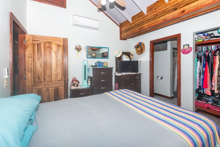 A cozy bedroom in Roatan with white walls, a wooden ceiling, and colorful striped bedding, featuring a wardrobe, small dresser, wall-mounted mirror, and hanging hats and accessories, set against a backdrop of natural wood accents and tropical decor in Honduras.