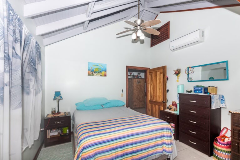 A cozy bedroom with a vaulted ceiling, white wooden beams, a ceiling fan, air conditioning unit, and light-colored walls in a vacation rental on Roatan, Honduras. The room features a bed with colorful striped bedding, a small bedside table, and various decorative elements including a mirror, paintings, and personal items.