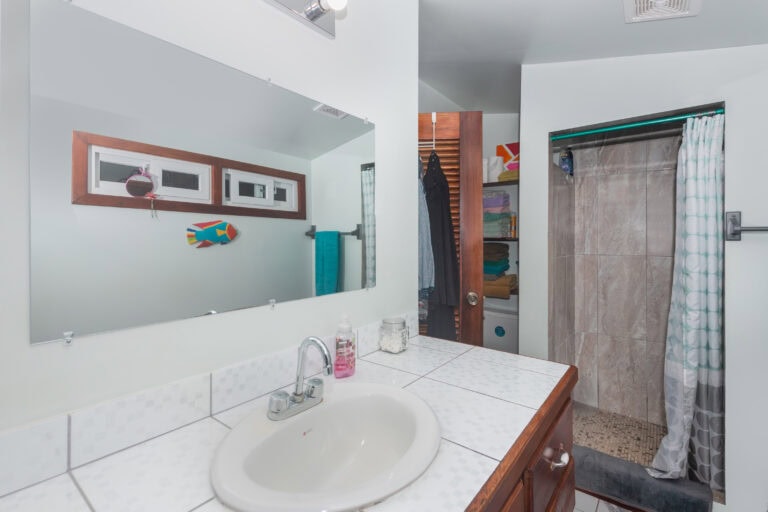 A bathroom with a white tiled countertop and sink, a large mirror above, and a small window with wooden trim on the wall; a storage shelf with folded towels and a sliding shower door with a curtain, all set within a setting that could be in a home or rental property in Roatan, Honduras.