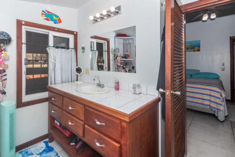 A cozy bathroom with a white tiled sink countertop, wooden cabinetry, and a large mirror, overlooking a bedroom with a colorful striped bedspread in a Roatan island home in Honduras.