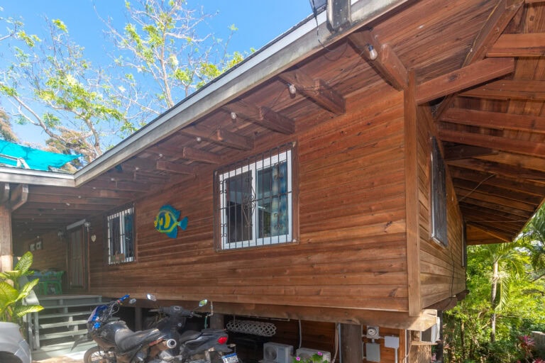 Wooden house with barred windows and a colorful fish decoration in Roatan, Bay Islands, Honduras, with green tropical plants and a clear blue sky in the background.