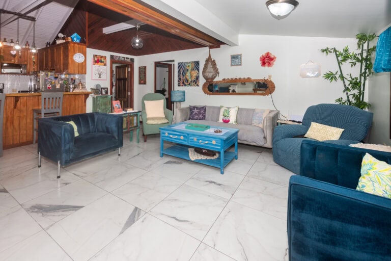 Living room with white walls, colorful decor, and tropical-themed artwork in a Roatan, Honduras home. The space features a blue coffee table, various comfortable chairs, and decorative pillows, creating a cozy atmosphere typical of Caribbean interiors.