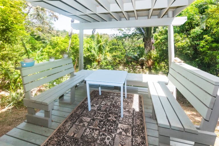 A white wooden outdoor seating area with benches and a table surrounded by lush green trees and tropical plants on Roatan, Honduras.