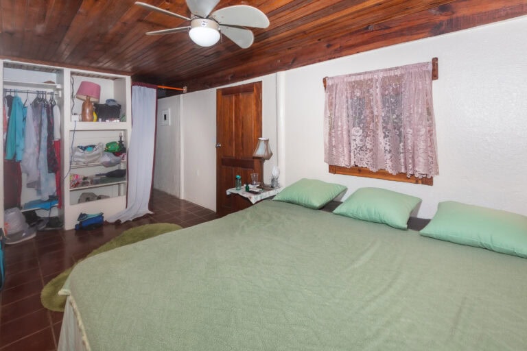 Interior view of a bedroom in Roatan, Honduras, featuring a large bed with green bedding and pillows, a wooden ceiling with a ceiling fan, a window with pink lace curtains, a small bedside table with decorative items, and an open closet with hanging clothes and shelves.