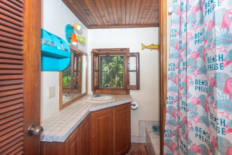 A rustic bathroom in Roatan, Honduras featuring a wooden ceiling, a small window with greenery outside, and a countertop with a sink, mirror, and colorful beach-themed curtains with starfish and shell patterns.