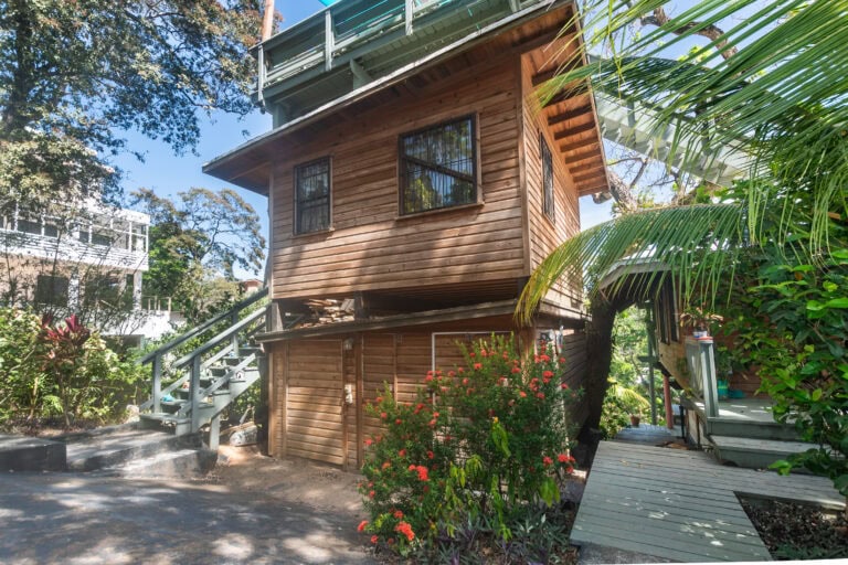 A wooden house with a green balcony on the second floor is surrounded by lush green trees and colorful flowering plants in Roatan, Honduras.